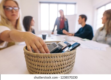 Female Putting Her Cell Phone In A Basket While Attending A Board Room Meeting In Her Office. No Cellphone Zone At Workplace Meeting.