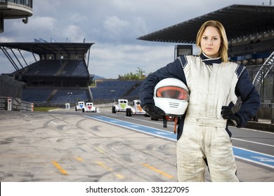 Female Race Car Driver, Holding Her Helmet Under Her Arm, Standing In The Pits Lane Of A Race Track Circuit