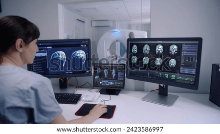 Female radiologist sits in control room and watches at monitors with displayed brain scans results. Professional doctor controls MRI or CT or PET scan with patient undergoing procedure at background.