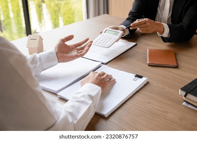 Female real estate agent in suit showing numbers and calculating house, property price on calculator for buyer or client while sitting at desk to discuss and negotiate about contract at office. - Shutterstock ID 2320876757