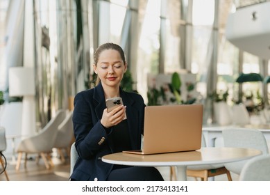 A female realtor in a blazer is reading news on the screen of a cell phone. A woman is sitting at the table in the modern lobby. A female IT developer near a laptop in a contemporary office interior. - Powered by Shutterstock