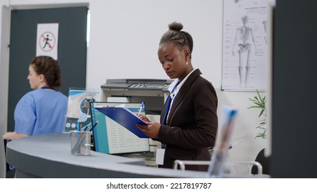 Female Receptionist Taking Notes On Papers And Using Medical Report To Prepare Consultation Appointments And Checkup Visits At Reception Counter. Hospital Worker At Registration Desk In Clinic Lobby.