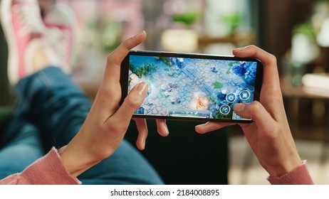 Female Is Relaxing On A Couch At Home, Playing An Interactive PvP RPG Strategy Video Game On Her Smartphone. Gamer Lies On A Sofa In Living Room. Close Up POV Photo Of Mobile Device Screen.