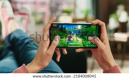 Female is Relaxing on a Couch at Home, Playing an Interactive PvP Shooter Video Game on Her Smartphone. Gamer Lies on a Sofa in Living Room. Close Up POV Photo of Mobile Device Screen.