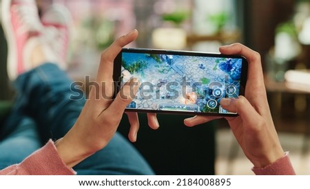Female is Relaxing on a Couch at Home, Playing an Interactive PvP RPG Strategy Video Game on Her Smartphone. Gamer Lies on a Sofa in Living Room. Close Up POV Photo of Mobile Device Screen.