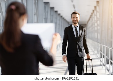 Female Representative Meeting Business Partner In Airport, Holding Placard With His Name. Handsome Businessman With Luggage Arrived For Business Meeting, Reading Names On Boards And Smiling