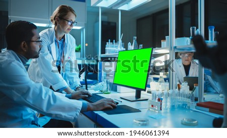 Female Research Scientist Has a Conversation with Bioengineer Next to a Desktop Computer with Green Screen Mock Up. They Look at Computer Template in a Modern Science Laboratory.