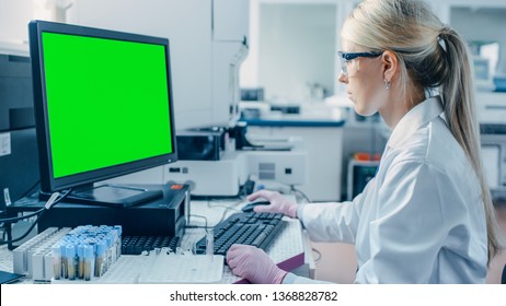 Female Research Scientist Sits At His Workplace In Laboratory, Uses Green Mock-up Screen Personal Computer. In The Background Genetics, Pharmaceutical Research Centre.