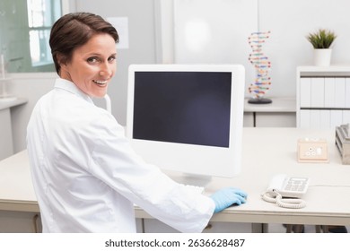Female researcher wearing lab coat and gloves sitting at lab bench smiling by computer DNA model. Scientist, research, laboratory, analysis, innovation, scientific, technology