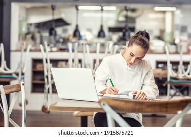 Female restaurant chef sits at the table after hours doing her bookkeeping in front of an open laptop.