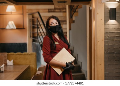 A Female Restaurant Manager In Black Disposable Medical Gloves Wears A Medical Face Mask Is Posing Holding A Wooden Menu In A Restaurant. A Cafe Owner With Long Hair In A Ruby Dress.