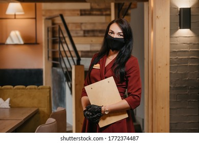 A Female Restaurant Manager In Black Disposable Medical Gloves Wears A Medical Face Mask Is Posing Holding A Wooden Menu In A Restaurant. A Charming Cafe Owner With Long Hair In A Ruby Dress.