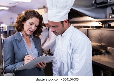 Female restaurant manager writing on clipboard while interacting to head chef in commercial kitchen - Powered by Shutterstock