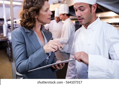 Female restaurant manager writing on clipboard while interacting to head chef in commercial kitchen - Powered by Shutterstock