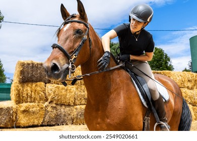 A female rider adjusts the reins of her brown horse while sitting in the saddle near a stack of hay bales. - Powered by Shutterstock