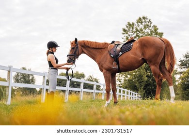 Female rider in equestrian clothes holding the reins and leading her beautiful saddled chestnut horse. Horseback riding activity concept. - Powered by Shutterstock