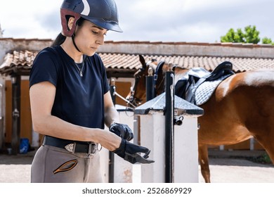 A female rider prepares for a ride by putting on gloves near a saddled brown horse at a stable, focusing on safety and readiness in an outdoor setting. - Powered by Shutterstock