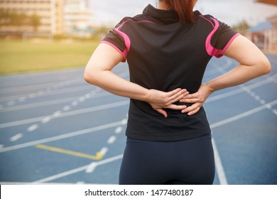 Female Runner Athlete Back Injury And Pain. Woman Suffering From Painful Lumbago While Running On The Blue Rubberized Running Track.