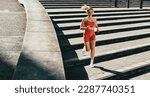 Female runner burning calories with a cardio workout. Sporty woman jogging down the stairs outdoors. Woman exercising with headphones on in the morning.