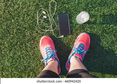 Female Runner Looking Down At Her Feet, Phone And Water Bottle In A Field