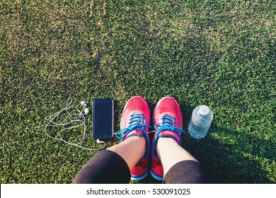 Female Runner Looking Down At Her Feet, Phone And Water Bottle In A Field