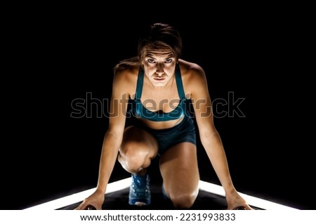 Female runner in race  start position. Girl in sportswear posing on lit track against dark background.
