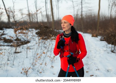 Female Runner Relaxing In Woods After Trail Running In Winter. Woman Wearing Hydration Vest Taking Break From Trail Running In Woods And Sipping Drink From Sports Bottle On Cold Winter Day.