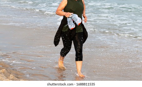 A Female Runner Is Running In The Water At The Edge Of The Beach Carrying Her Shoes In Her Hands To Keep Them Dry During One Mile Race.