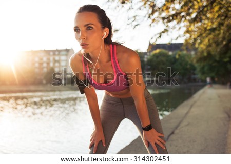 Female runner standing bent over and catching her breath after a running session along lake in city. Young sports woman taking break after a run.