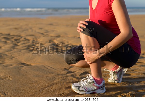 Female runner tibia muscle cramp. Woman\
clutching her shin because of a running injury and inflammation.\
Tibial periostitis hurt while jogging on\
beach.