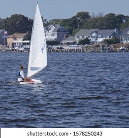 Female Sailing A Sunfish Sailboat In The Bay With Large Homes And Trees In The Distance