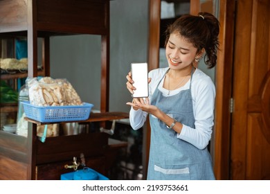 Female Salesperson In Apron Showing Phone Screen To Camera With Cart Background