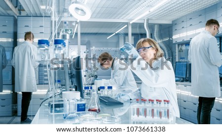 Female Scientist Analyzes Liquid in the Beaker and Types Down Observations on Her Computer. She's Working in a Busy Laboratory Full of Scientists Conducting Experiments.
