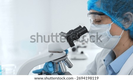 Female scientist analyzing sample through microscope, wearing protective lab gear during medical research investigation. Medicine, healthcare and science concept