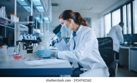 Female Scientist in Face Mask and Glasses Looking a Petri Dish with Genetically Modified Sample Chemicals Under a Microscope. Microbiologist Working in Modern Laboratory with Technological Equipment.