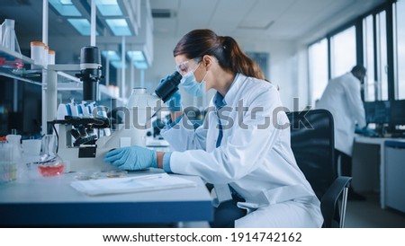 Female Scientist in Face Mask and Glasses Looking a Petri Dish with Genetically Modified Sample Chemicals Under a Microscope. Microbiologist Working in Modern Laboratory with Technological Equipment.