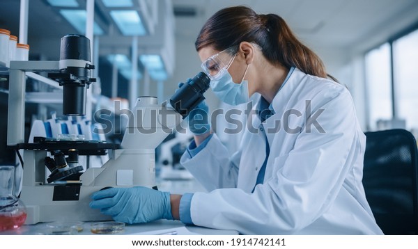 Female Scientist in Face Mask and Glasses\
Looking a Petri Dish with Genetically Modified Sample Chemicals\
Under a Microscope. Microbiologist Working in Modern Laboratory\
with Technological\
Equipment.
