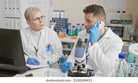 A female scientist and her male colleague in lab coats analyze samples in a laboratory while the man talks on the phone. - Powered by Shutterstock