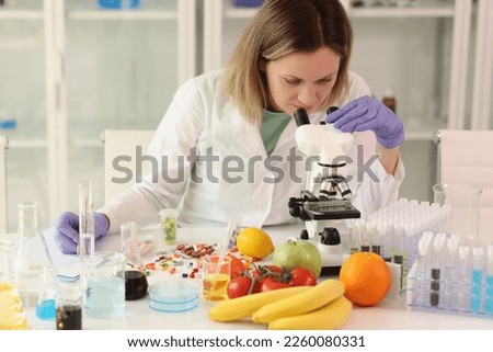 Female scientist with microscope, test tubes, laboratory glassware and reagents examining many fruits and vegetables in her lab. Dieting and food quality research concept.