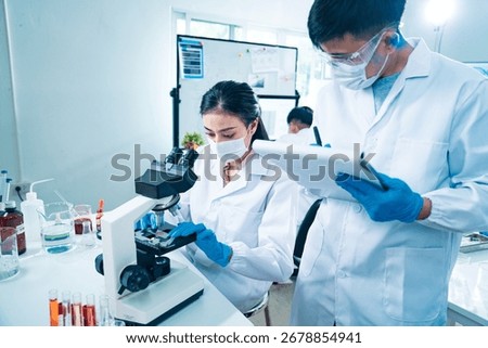 Female scientist using a microscope to examine a blood sample while a colleague records data, symbolizing teamwork, healthcare diagnostics, virus testing, and innovation in medical research.