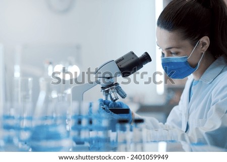 Female scientist working in the lab, she is wearing a surgical mask and using a microscope