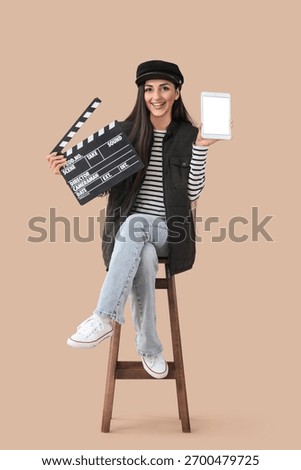 Female scriptwriter with blank tablet computer and movie clapper sitting on beige background