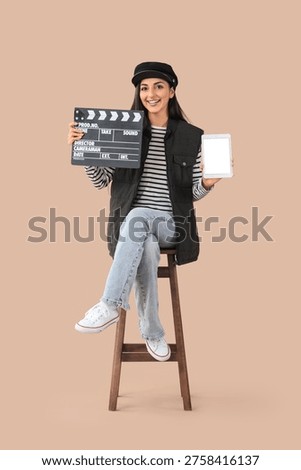 Female scriptwriter with blank tablet computer and movie clapper sitting on beige background