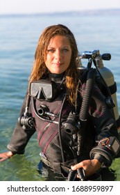 Female Scuba Diving Instructor Standing In Water Wearing A Dry Suit, A Twin Tank And Holding Fins