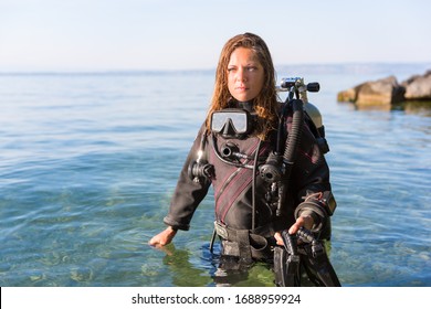 Female Scuba Diving Instructor Standing In Water Wearing A Dry Suit, A Twin Tank And Holding Fins