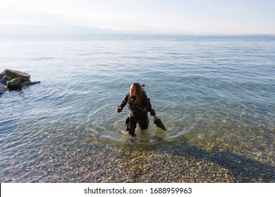 Female Scuba Diving Instructor Standing In Water Wearing A Dry Suit, A Twin Tank And Holding Fins