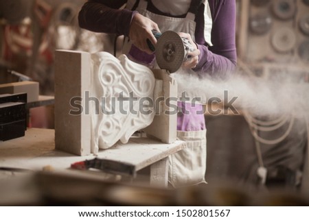 Female sculptor cutting a white marble piece with a power tool, craftswoman shaping a sculpture with an angle grinder, caucasian woman working inside an arts workshop, detail closeup of hands and dust