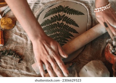 Female sculptor making clay pottery in a home workshop.Making an imprint of a fern leaf on a clay plate,hands close-up.Small business,entrepreneurship,hobby, leisure,sustainability concept. 