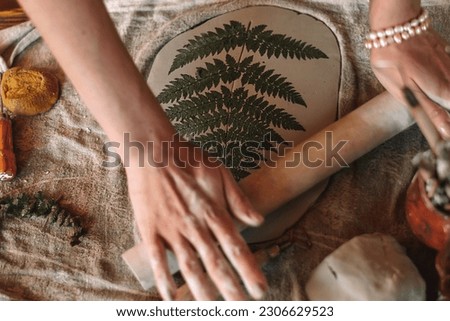 Female sculptor making clay pottery in a home workshop.Making an imprint of a fern leaf on a clay plate,hands close-up.Small business,entrepreneurship,hobby, leisure,sustainability concept. 