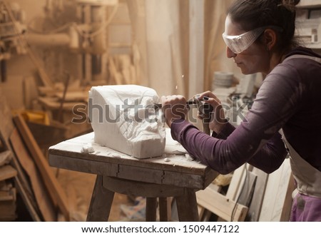 Female sculptor at work in a workshop, using hammer and chisel to sculpt a piece of white marble stone, debris and particles flying around, stonemasonry and stonecraft, copy space